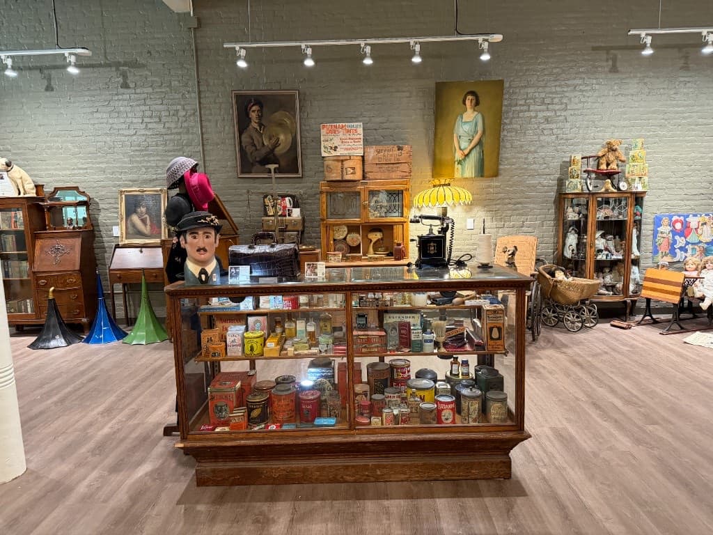 Historic general-store glass counter filled with tins, packages, toys, and household goods, with portraits behind.