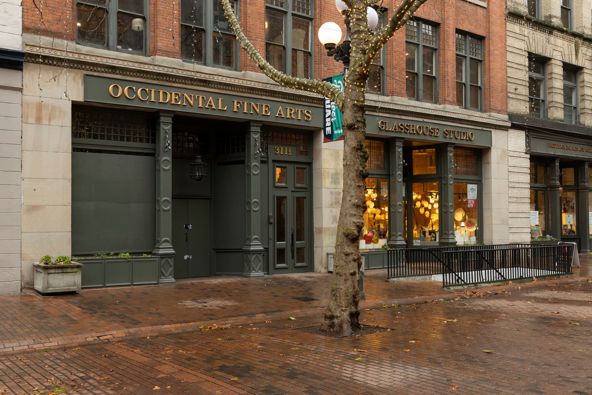 The Occidental Fine Arts Center storefront on Occidental Avenue at dusk, golden gallery light spilling onto wet brick.